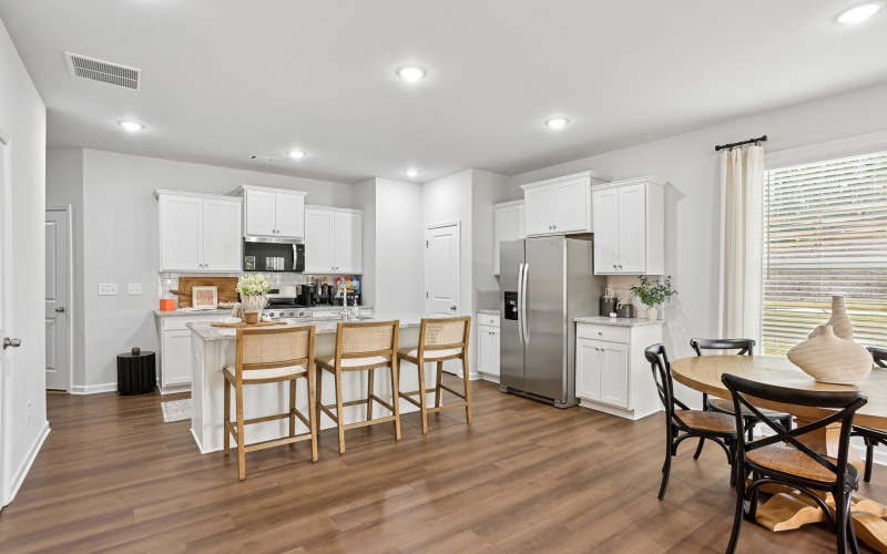 Pet-Friendly Townhomes In Summerville, SC - ABODE At Summers Corner - A Kitchen With White Shaker Cabinets, A Large Granite Island With Three Woven Barstools, And Stainless-Steel Appliances.